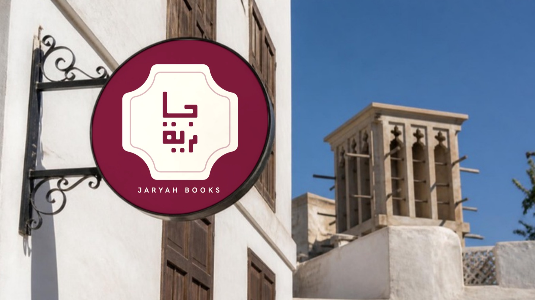 لافتة دائرية معلّقة عليها الاسم بالكوفي داخل إطارٍ مفصّص، في شارعٍ من بيوت الحجر المرجاني القديمة