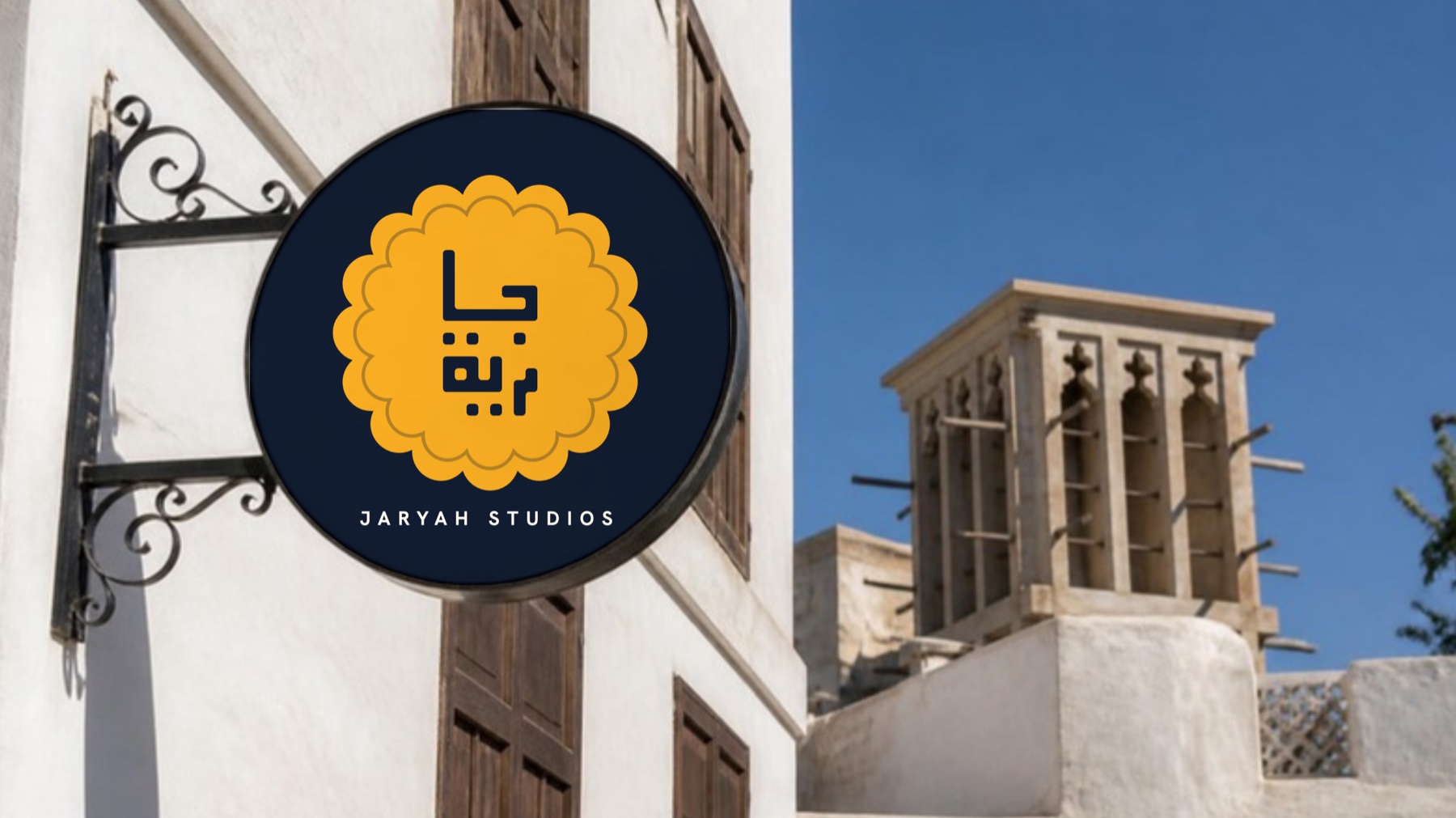 لافتة دائرية معلّقة عليها الشمسة العسلية وJaryah Studios، في شارعٍ من بيوت الحجر المرجاني وبرجٌ للهواء